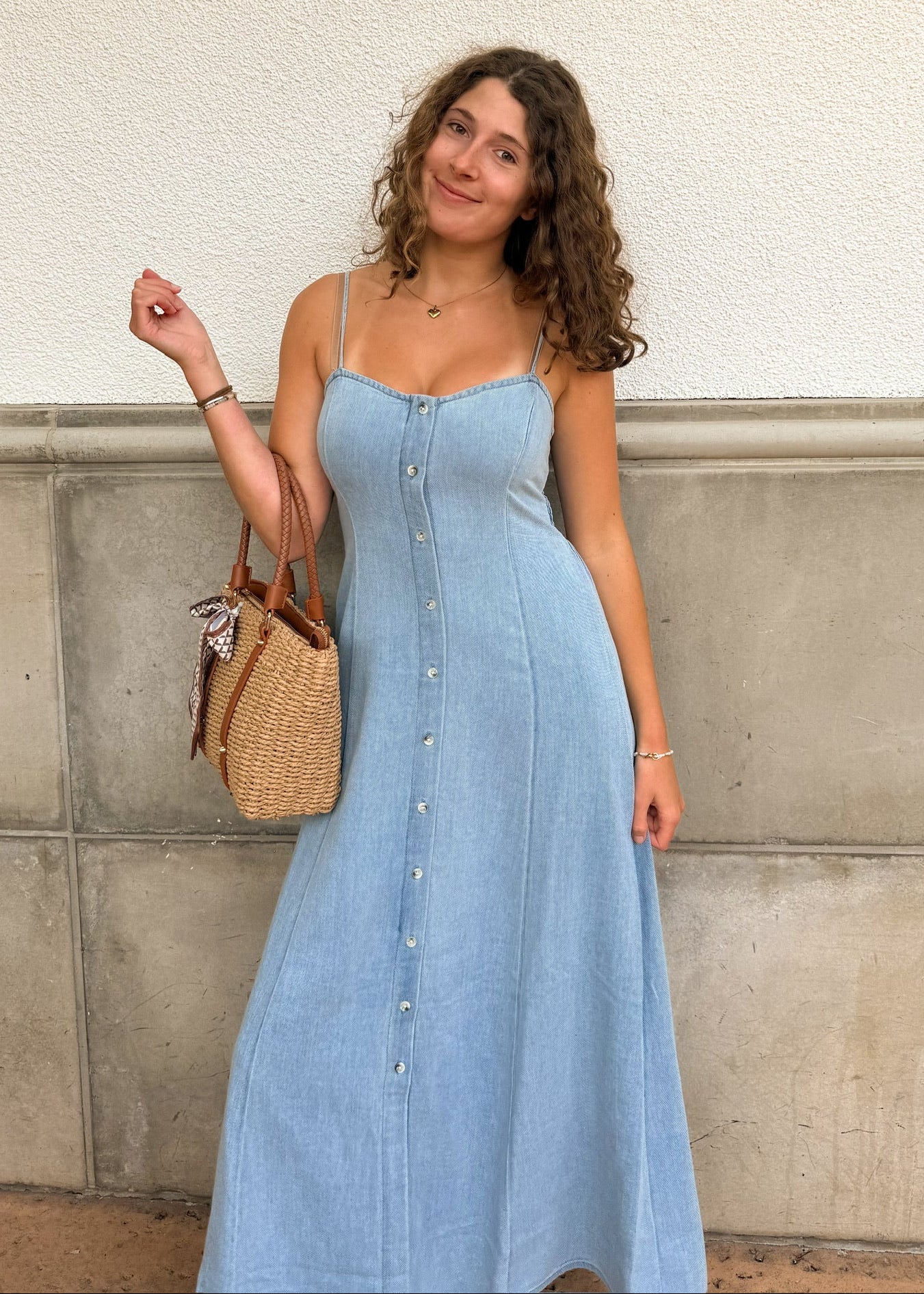 Woman in a light blue dress holding a woven bag against a concrete wall.